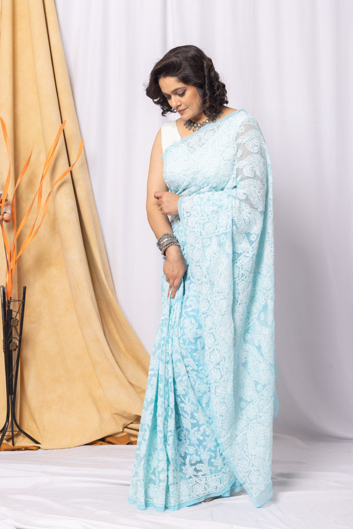 A woman posing in a sky blue georgette saree with allover chikankari hand embroidery, paired with a matching blouse, standing against a white backdrop.
