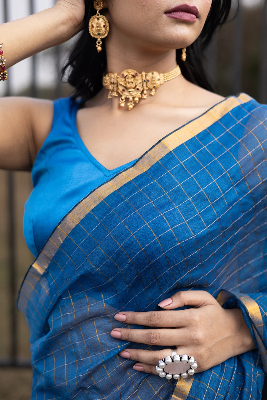 Woman in a blue saree standing outdoors with a metal fence and grassy area in the background.