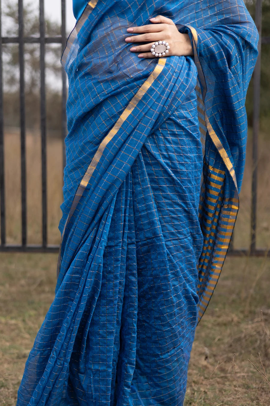 Woman in a blue saree standing outdoors with a metal fence and grassy area in the background.