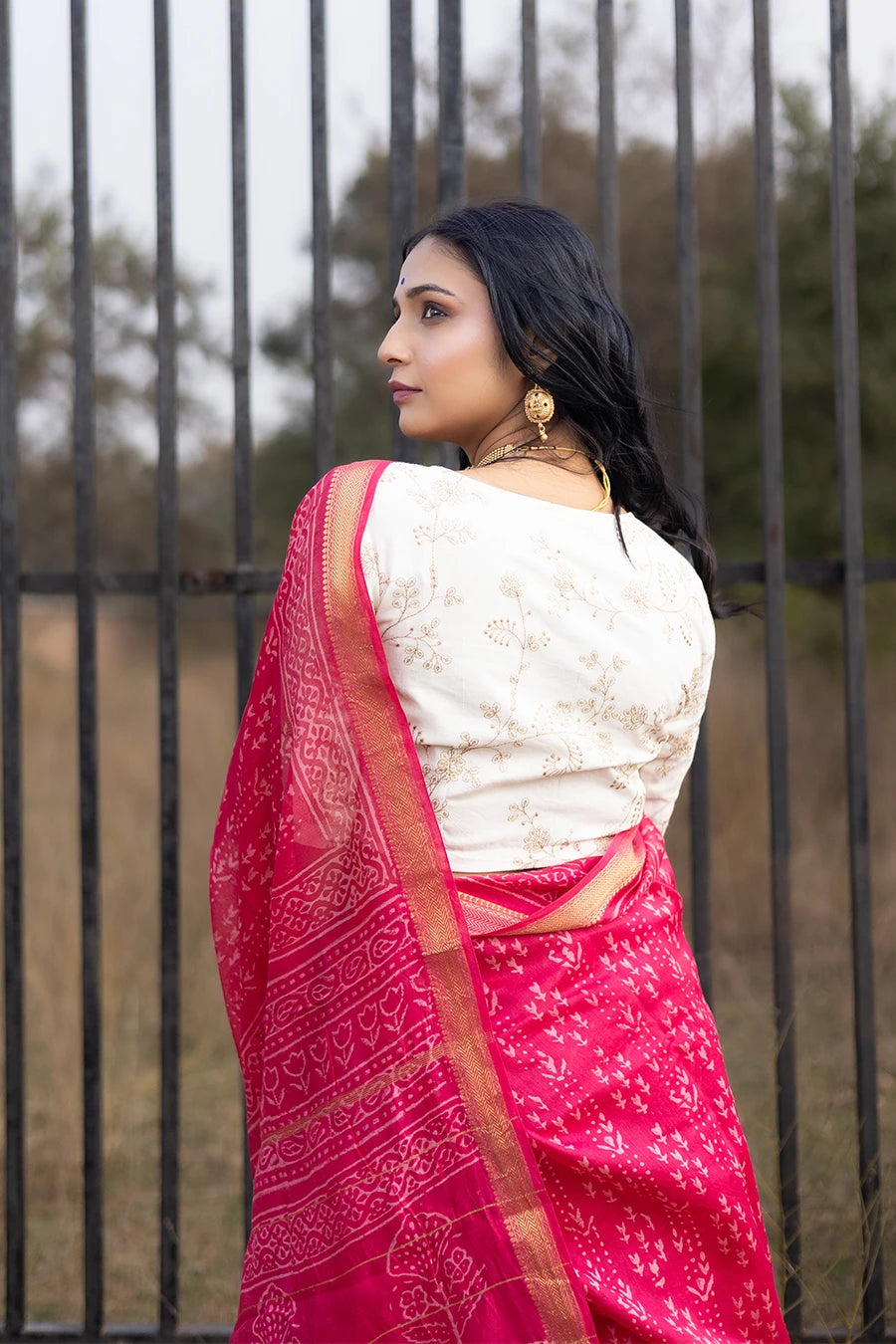 Woman in a red saree with white patterns standing outdoors near a metal fence.