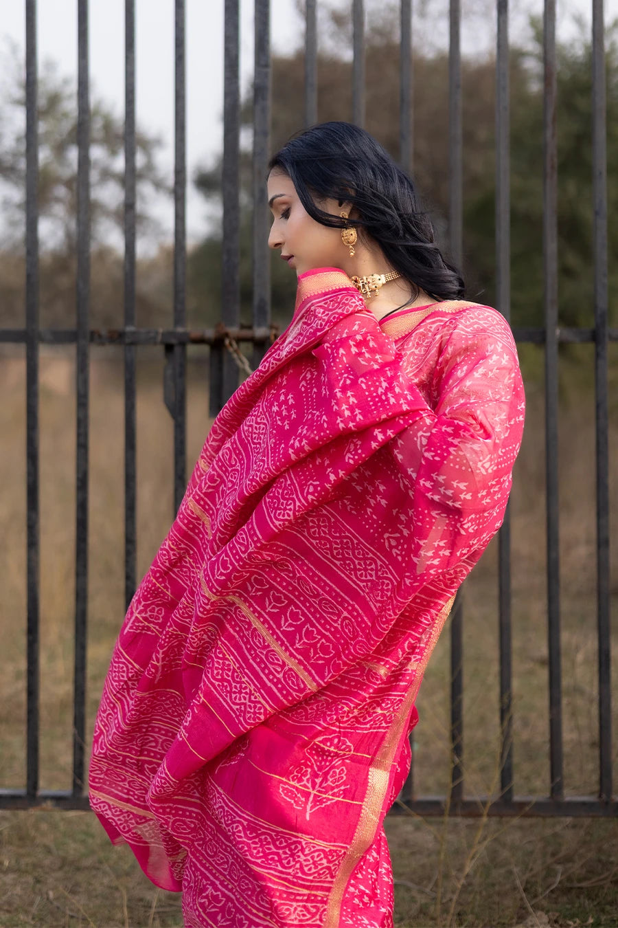 Woman in a red saree with white patterns standing outdoors near a metal fence.