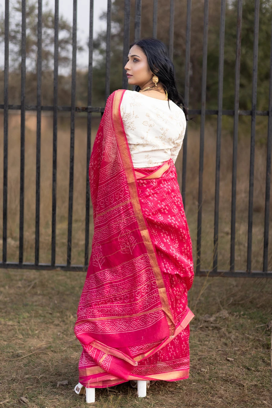 Woman in a red saree with white patterns standing outdoors near a metal fence.