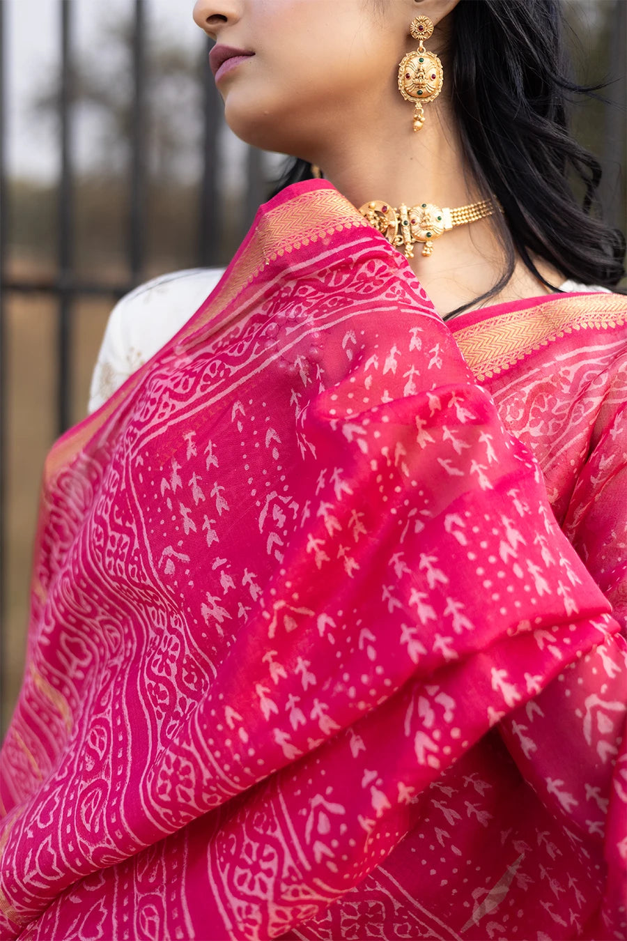Woman in a red saree with white patterns standing outdoors near a metal fence.