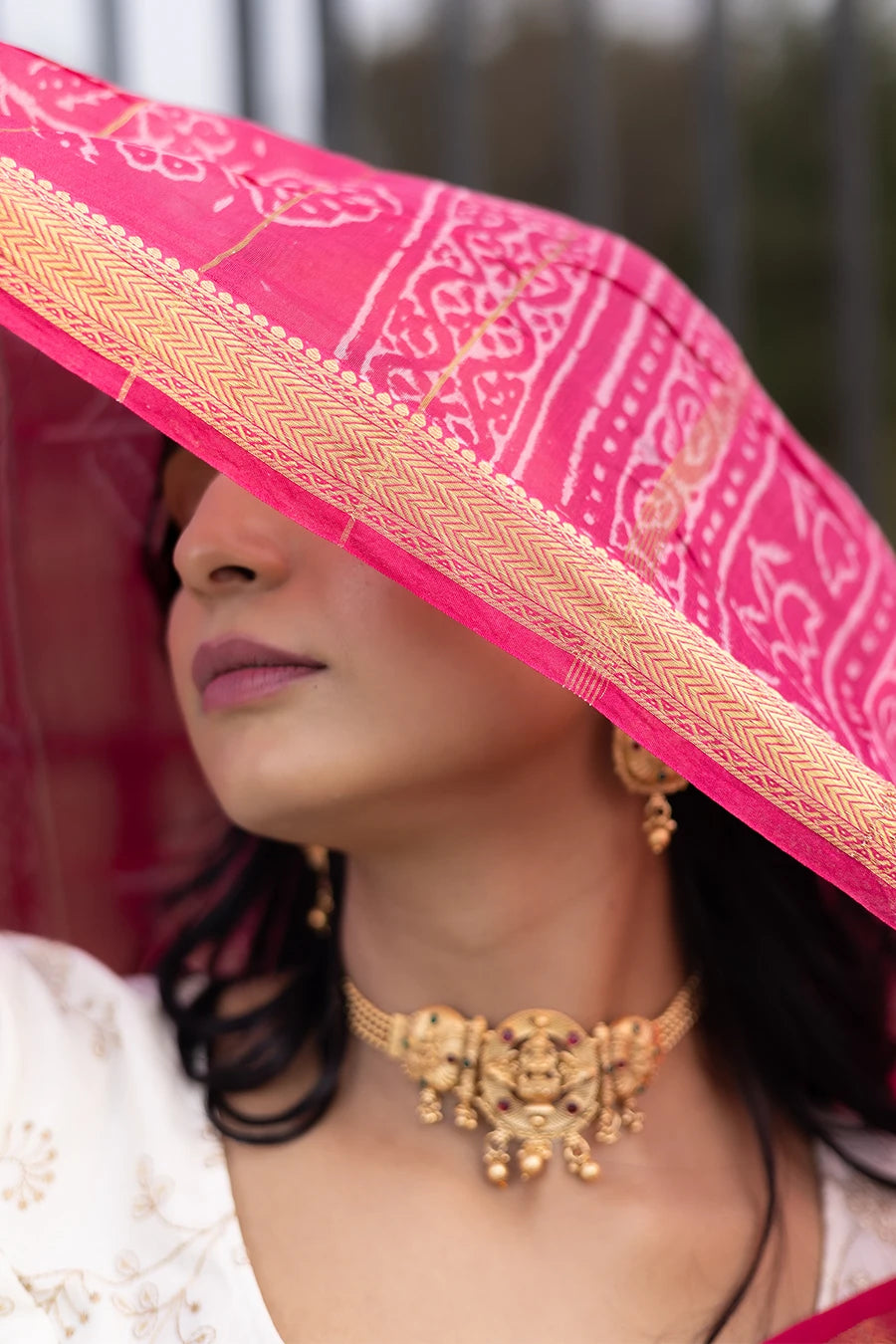 Woman in a red saree with white patterns standing outdoors near a metal fence.