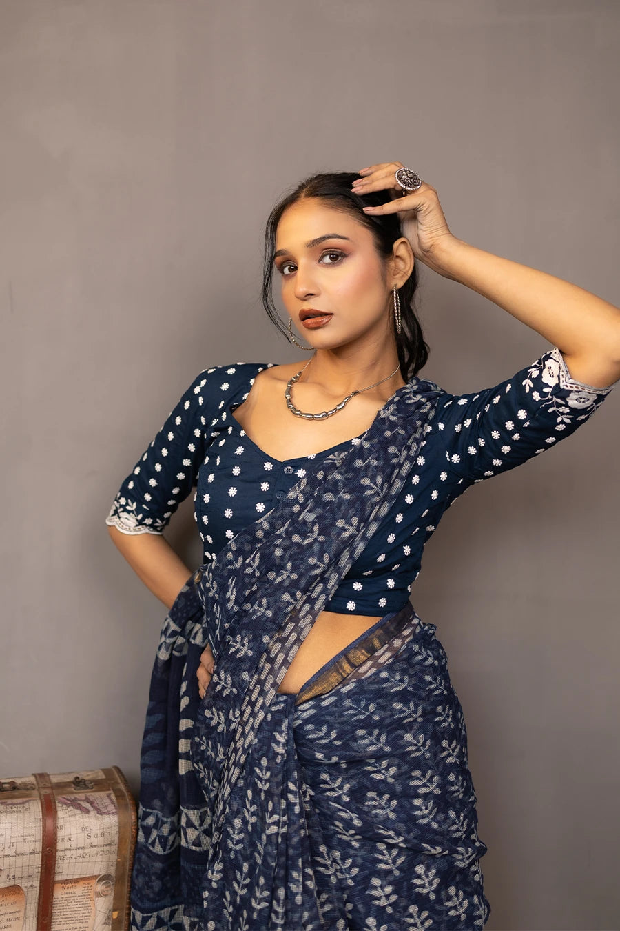 Woman in a patterned outfit standing next to a small table with books and a suitcase against a plain background