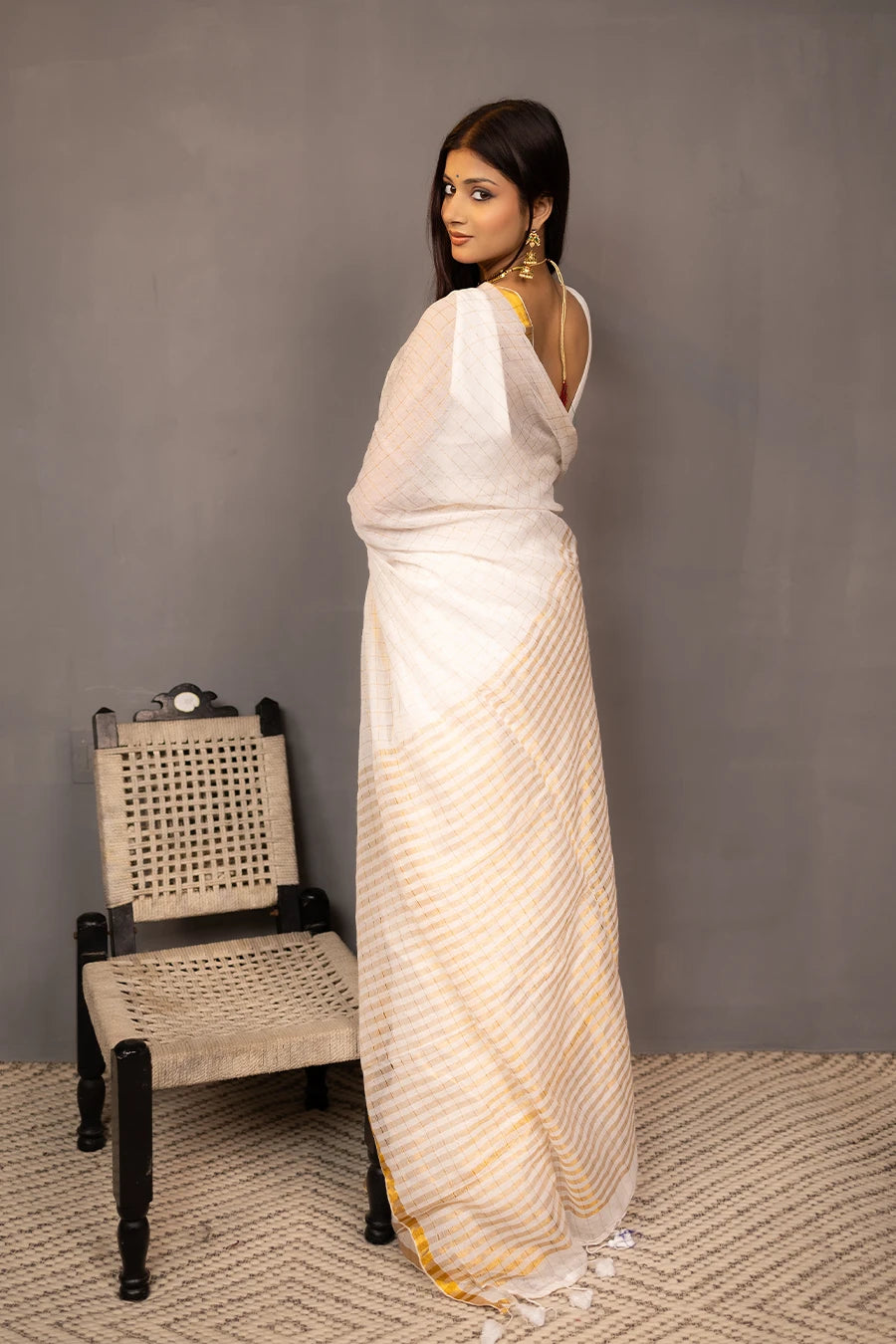 Woman in a white saree with gold border standing next to a wooden chair against a grey wall.