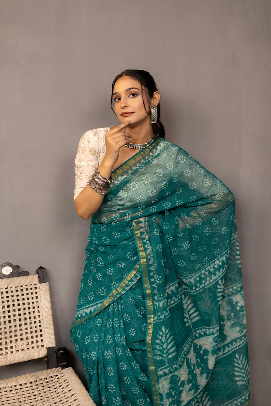 Woman in a teal saree standing next to a chair against a grey wall.