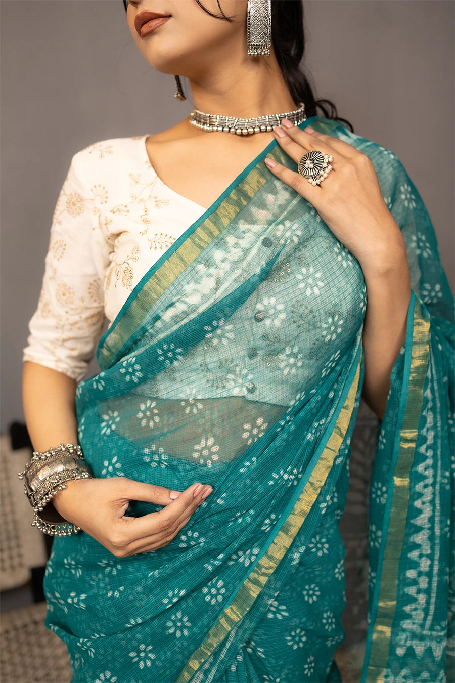 Woman in a teal saree standing next to a chair against a grey wall.