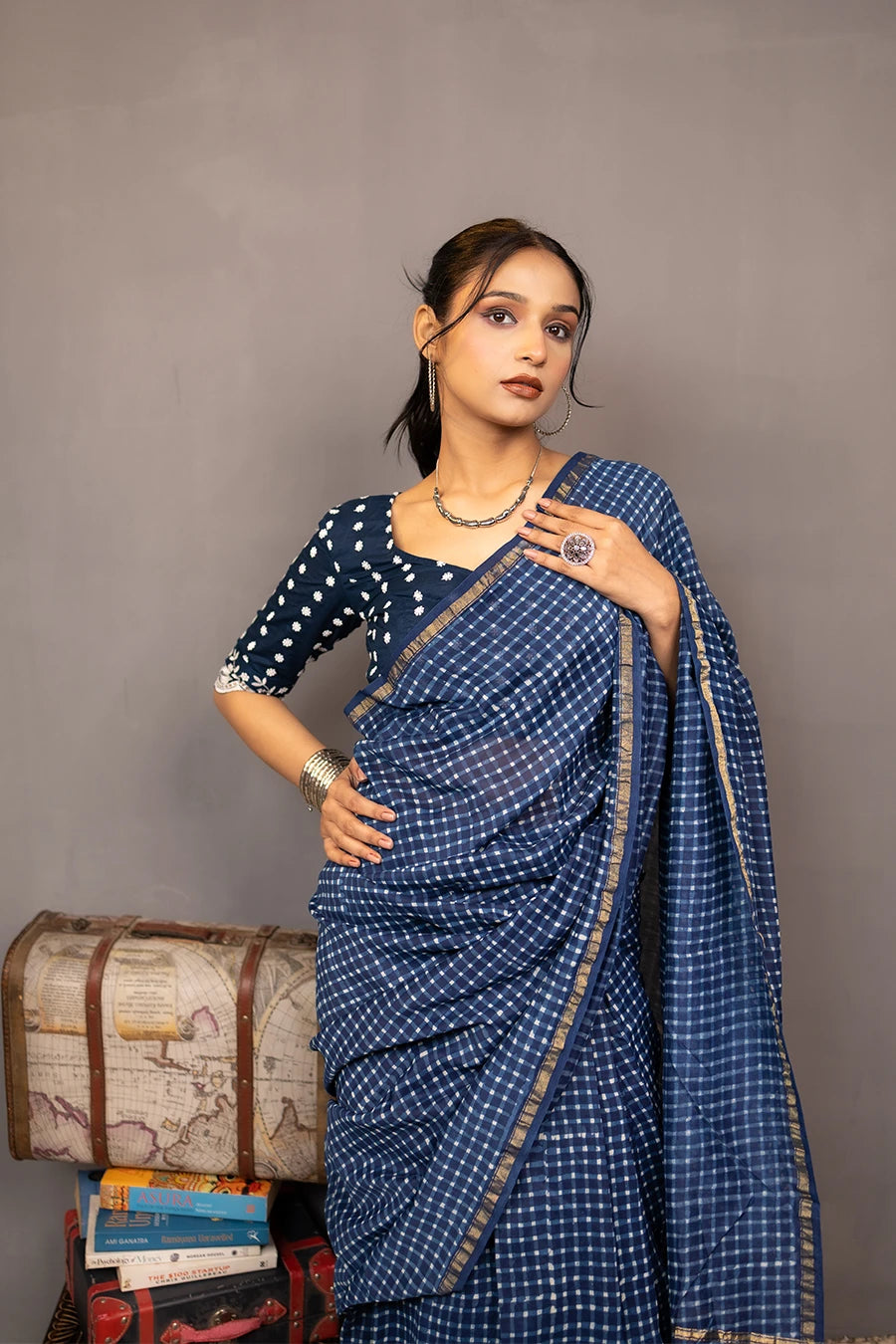 Woman in a blue saree standing next to a small wooden table with books and a suitcase against a grey background.
