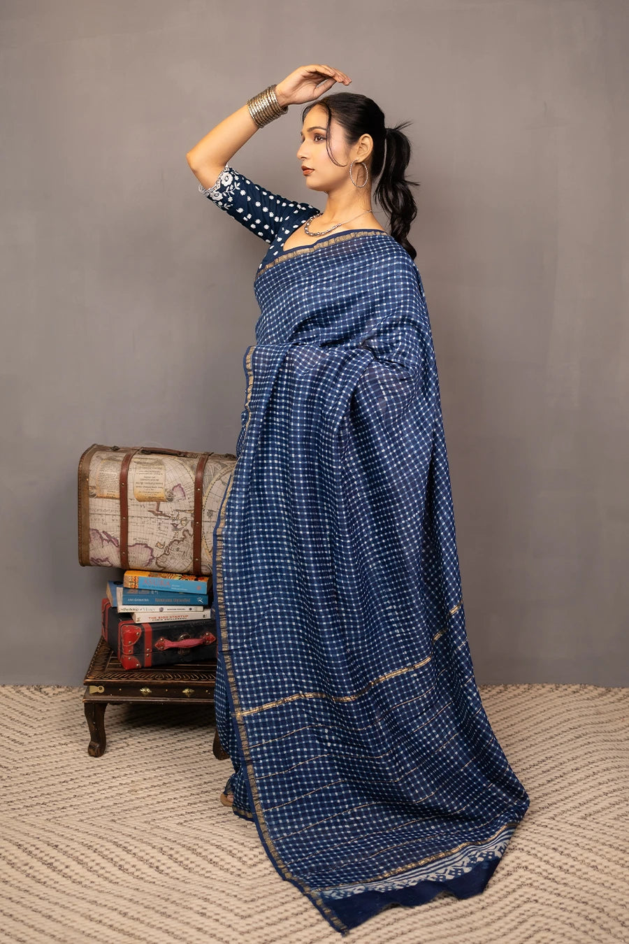 Woman in a blue saree standing next to a small wooden table with books and a suitcase against a grey background.