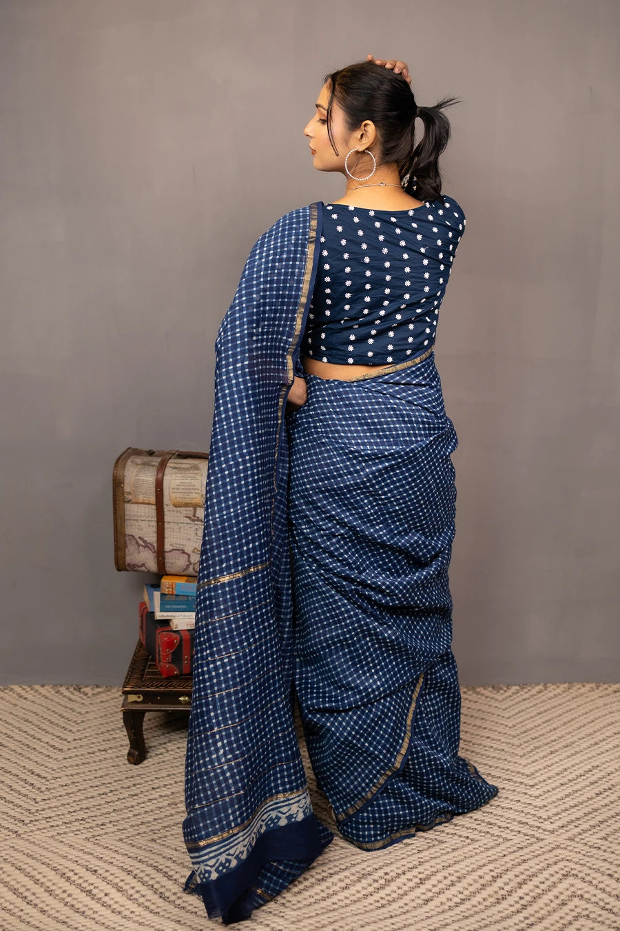 Woman in a blue saree standing next to a small wooden table with books and a suitcase against a grey background.