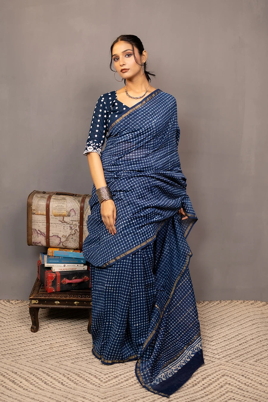 Woman in a blue saree standing next to a small wooden table with books and a suitcase against a grey background.