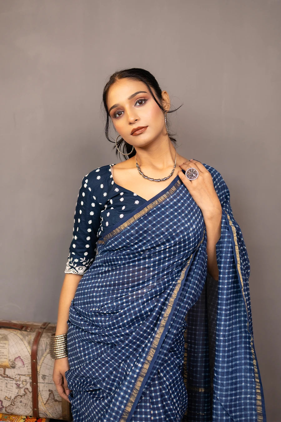 Woman in a blue saree standing next to a small wooden table with books and a suitcase against a grey background.