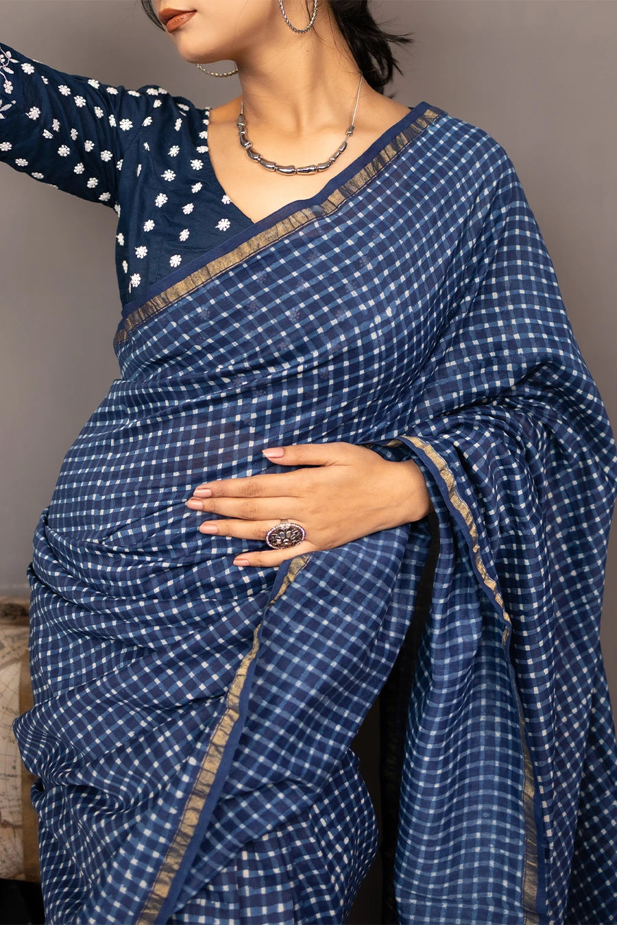 Woman in a blue saree standing next to a small wooden table with books and a suitcase against a grey background.
