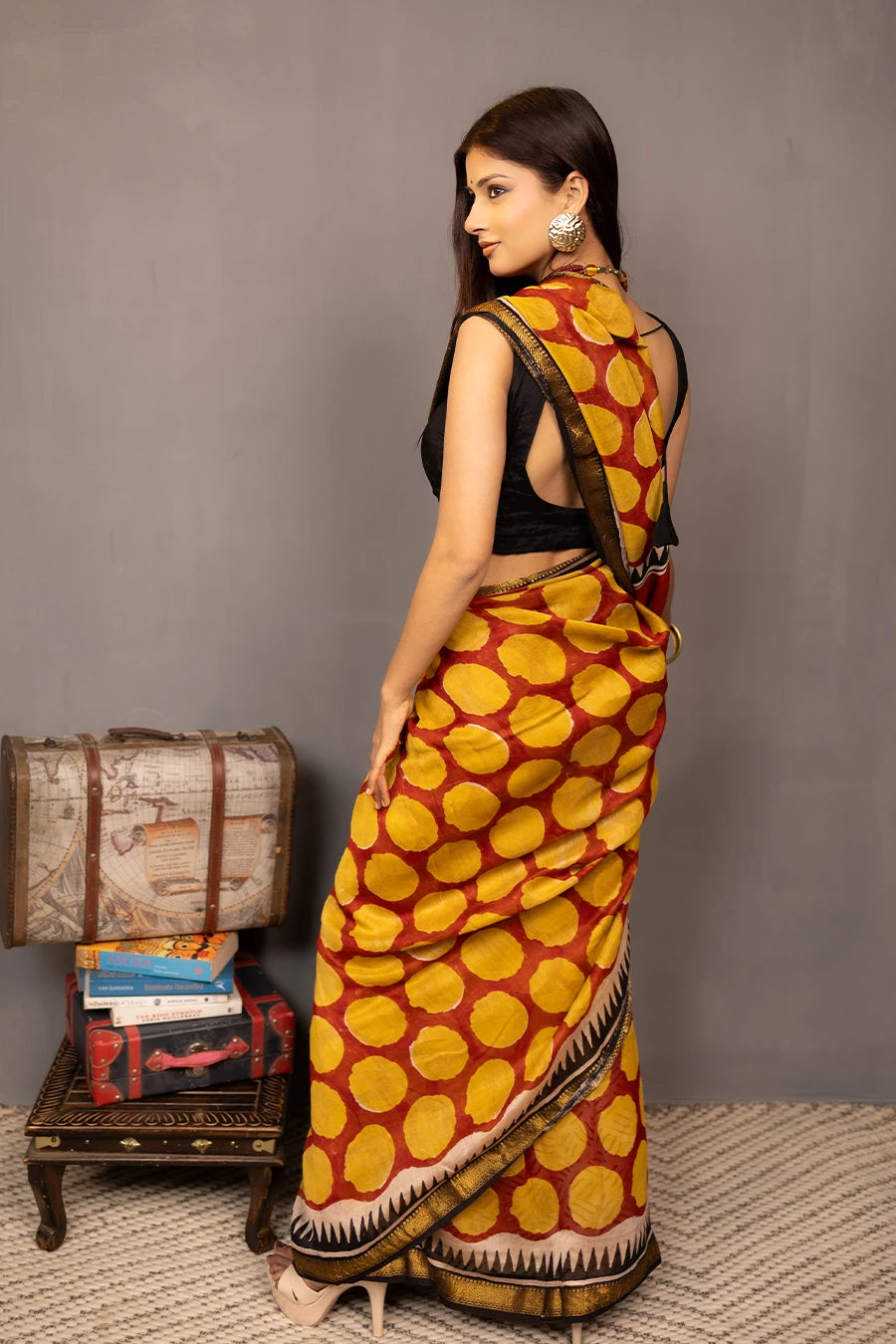 Woman wearing a traditional saree with a patterned design, standing next to a small table with books and a suitcase.