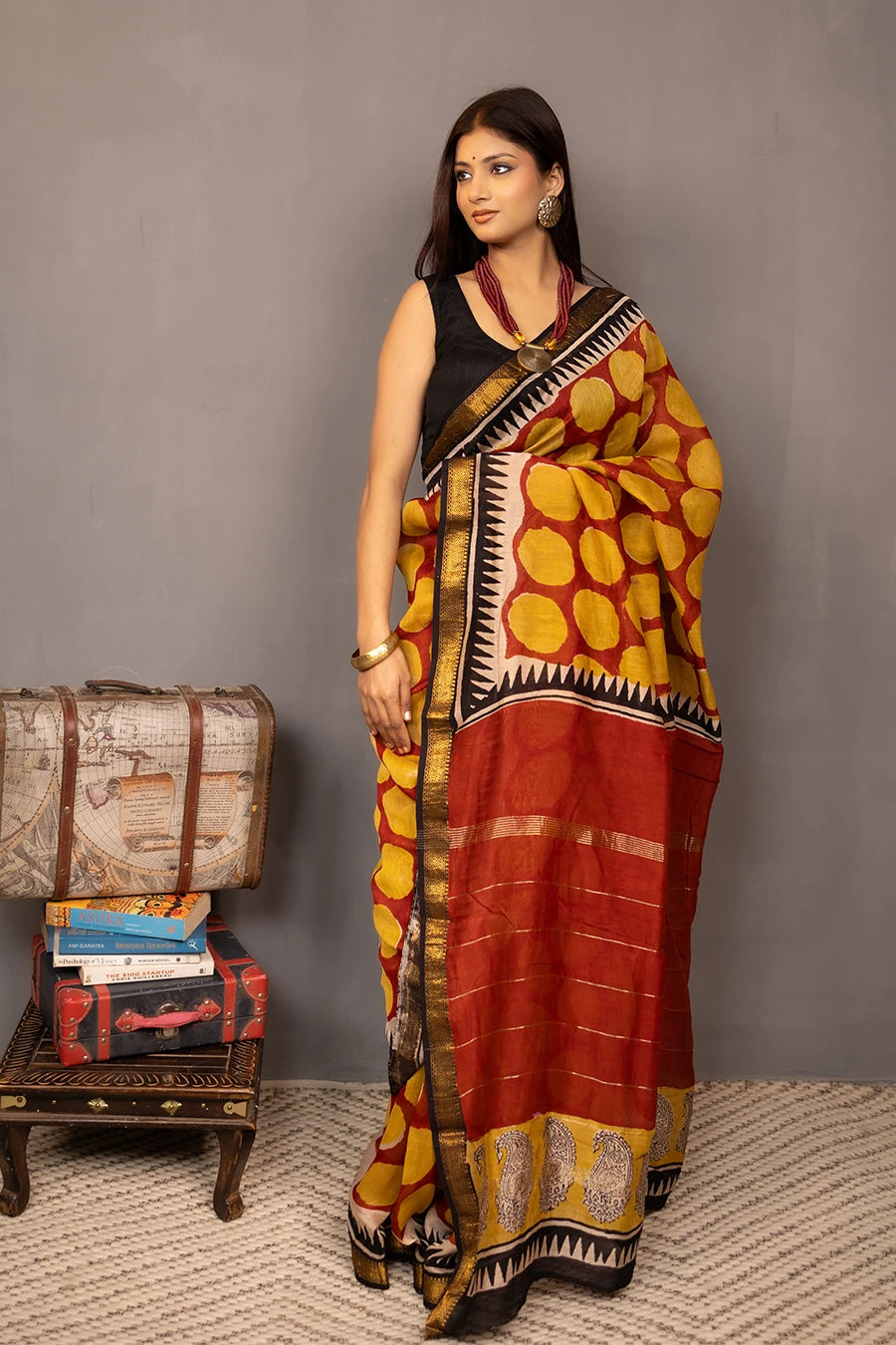 Woman wearing a traditional saree with a patterned design, standing next to a small table with books and a suitcase.