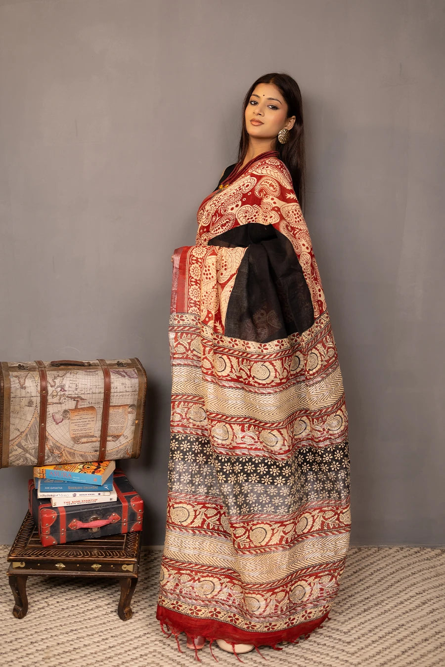 Woman wearing a traditional saree with a patterned design, standing next to a small table with books and a suitcase.