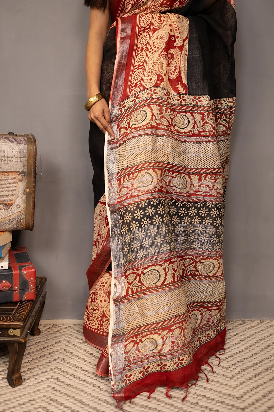 Woman wearing a traditional saree with a patterned design, standing next to a small table with books and a suitcase.