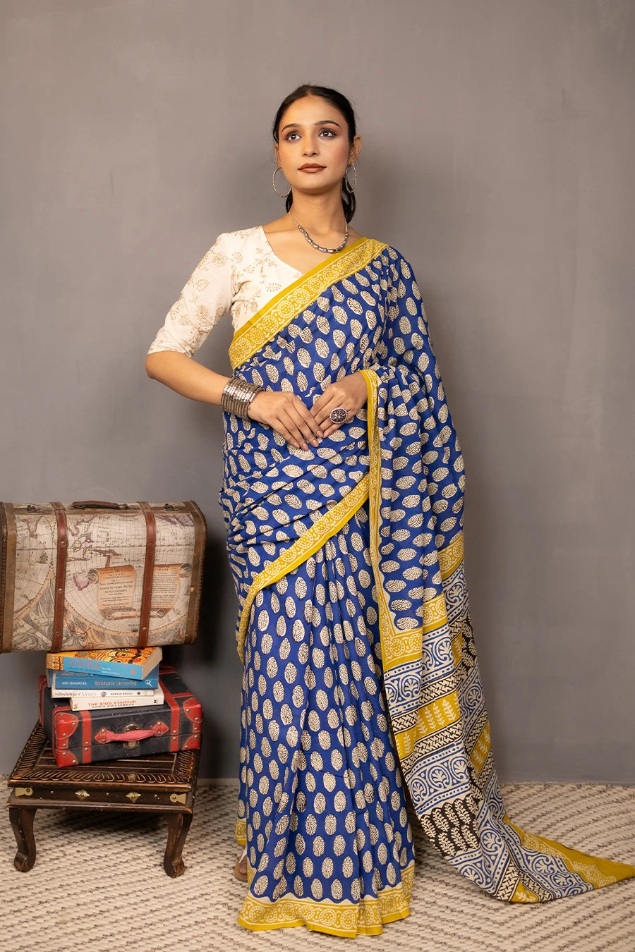 Woman wearing a blue and yellow saree standing next to vintage suitcases and books against a grey background