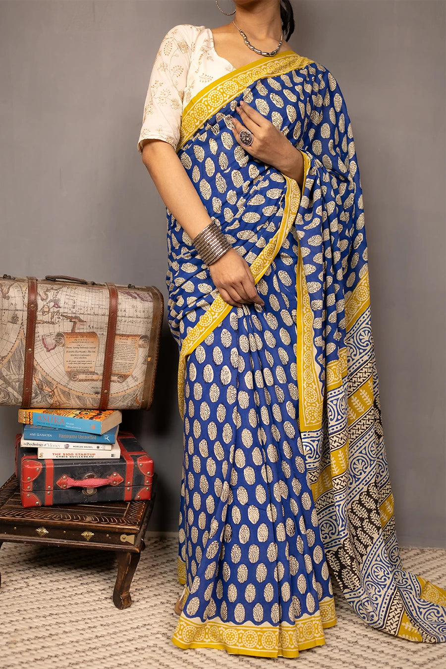 Woman wearing a blue and yellow saree standing next to vintage suitcases and books against a grey background