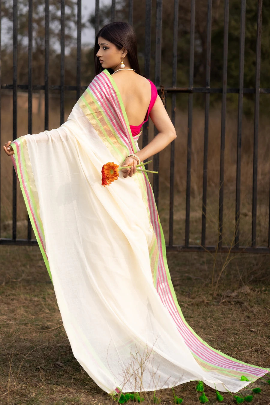 Model wearing an off-white saree with pink and green striped border and tassel detailing, lightweight and suitable for festive and casual occasions.