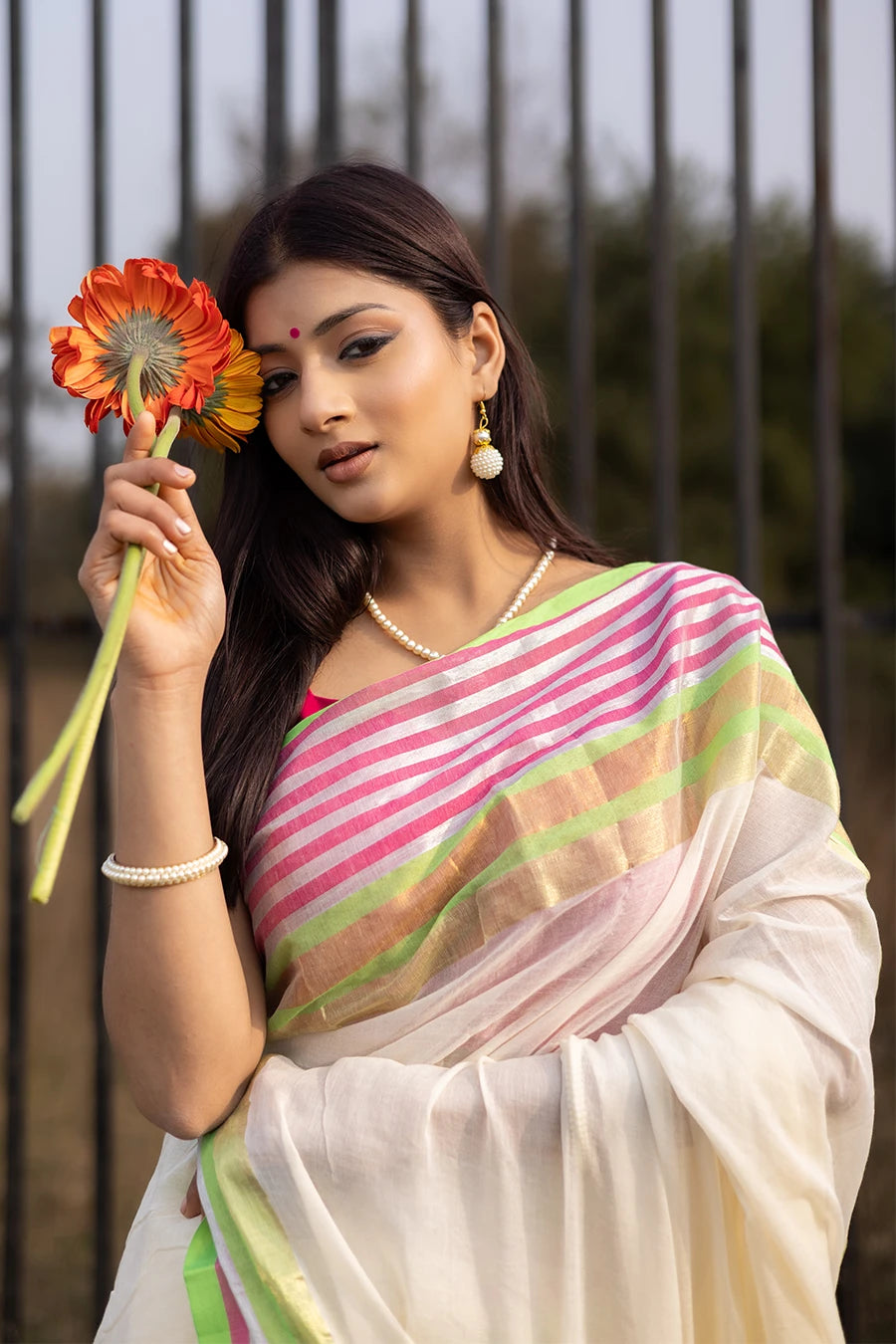 Model wearing an off-white saree with pink and green striped border and tassel detailing, lightweight and suitable for festive and casual occasions.