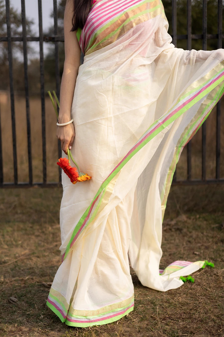 Model wearing an off-white saree with pink and green striped border and tassel detailing, lightweight and suitable for festive and casual occasions.