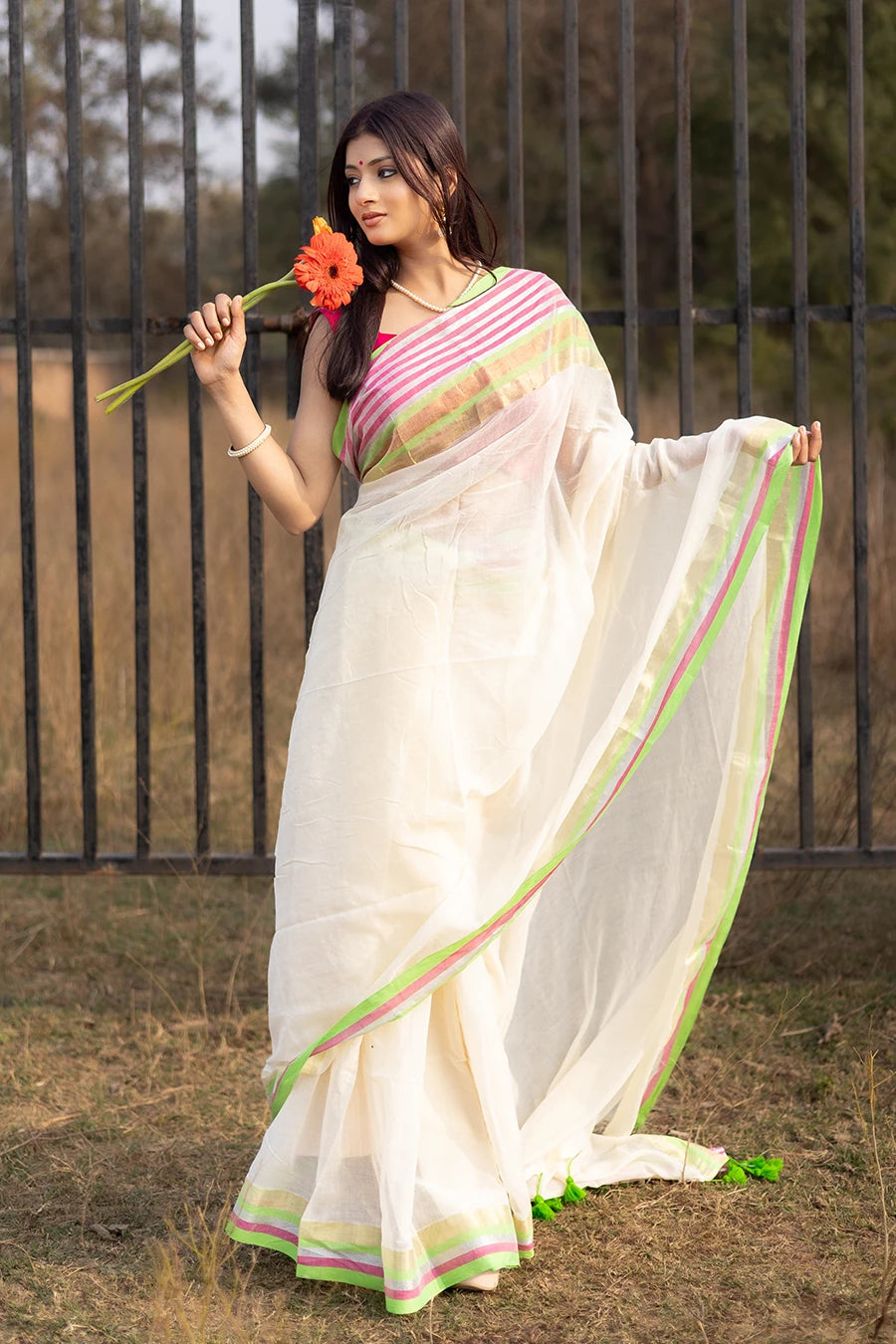 Model wearing an off-white saree with pink and green striped border and tassel detailing, lightweight and suitable for festive and casual occasions.