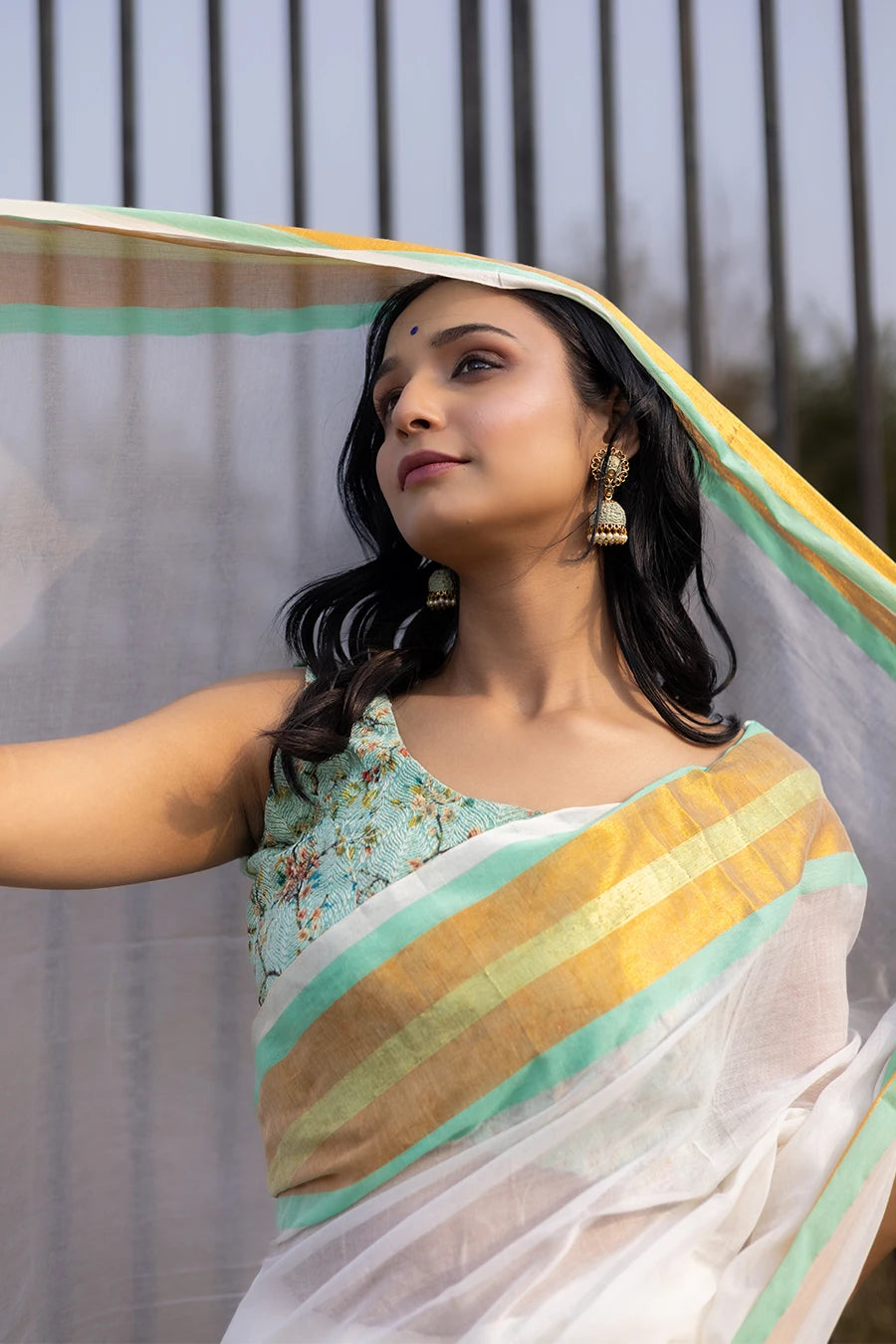 Model wearing an off-white saree with gold and mint green striped border and tassel detailing, lightweight and suitable for festive and casual occasions.