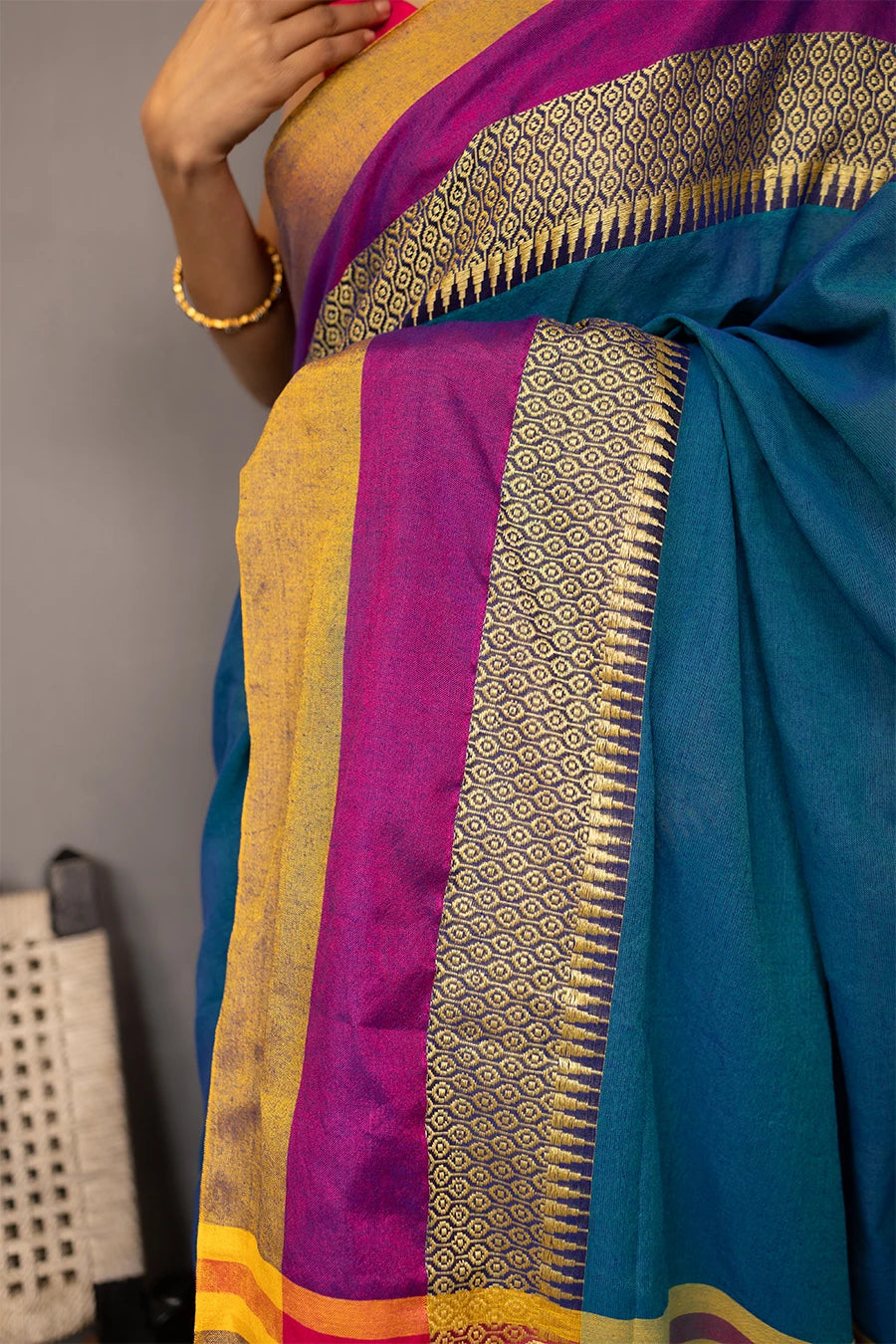 Woman wearing a blue saree with gold and purple accents, standing next to a chair against a grey background.