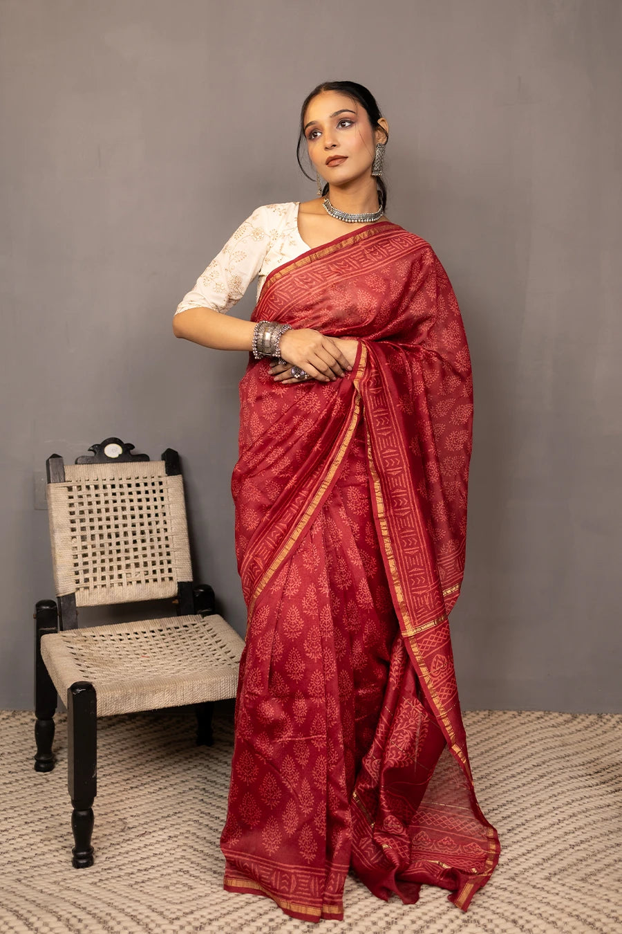 Woman in a red saree standing next to a chair against a grey wall.