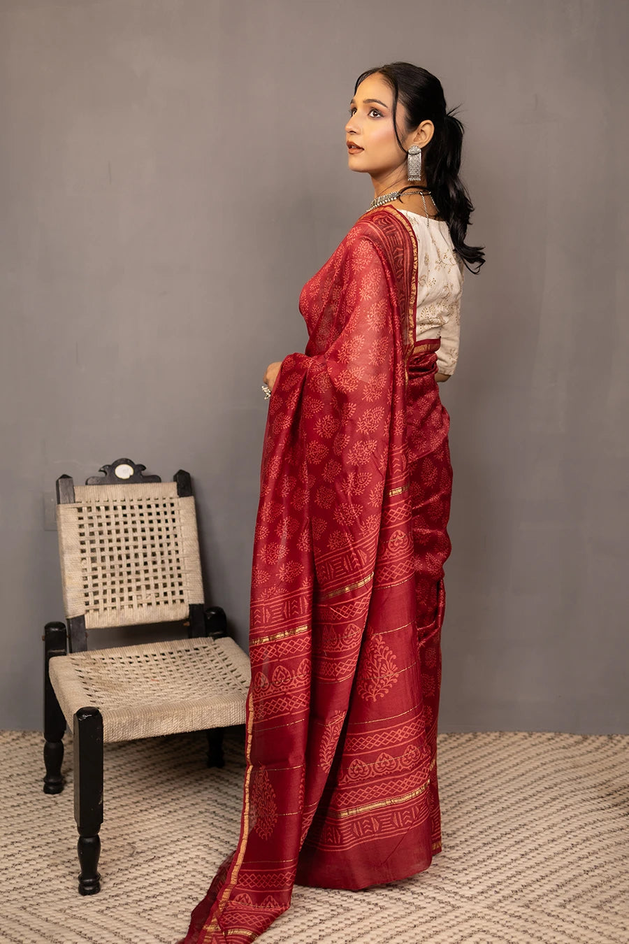 Woman in a red saree standing next to a chair against a grey wall.