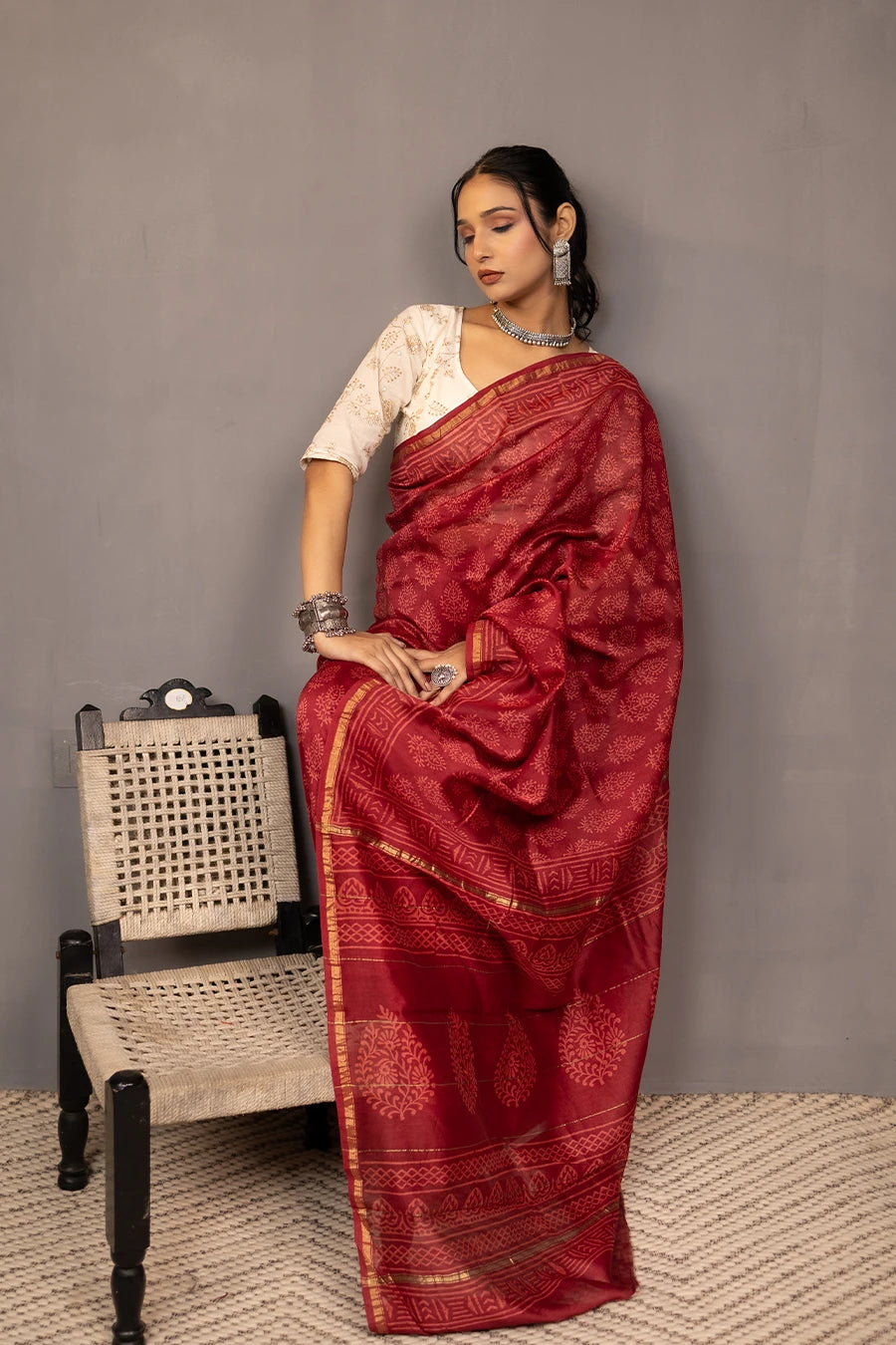 Woman in a red saree standing next to a chair against a grey wall.