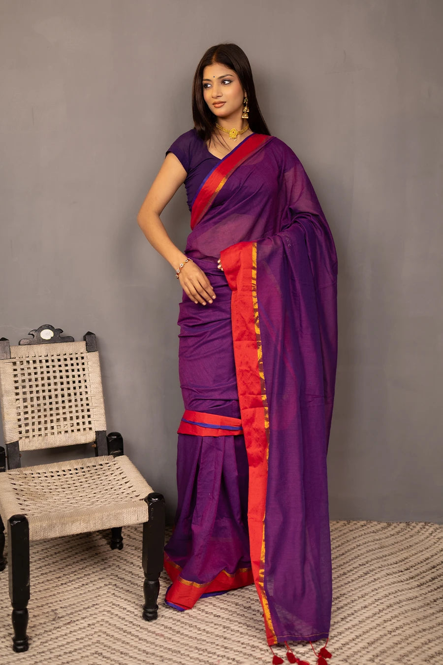Woman wearing a purple saree with red border against a grey background