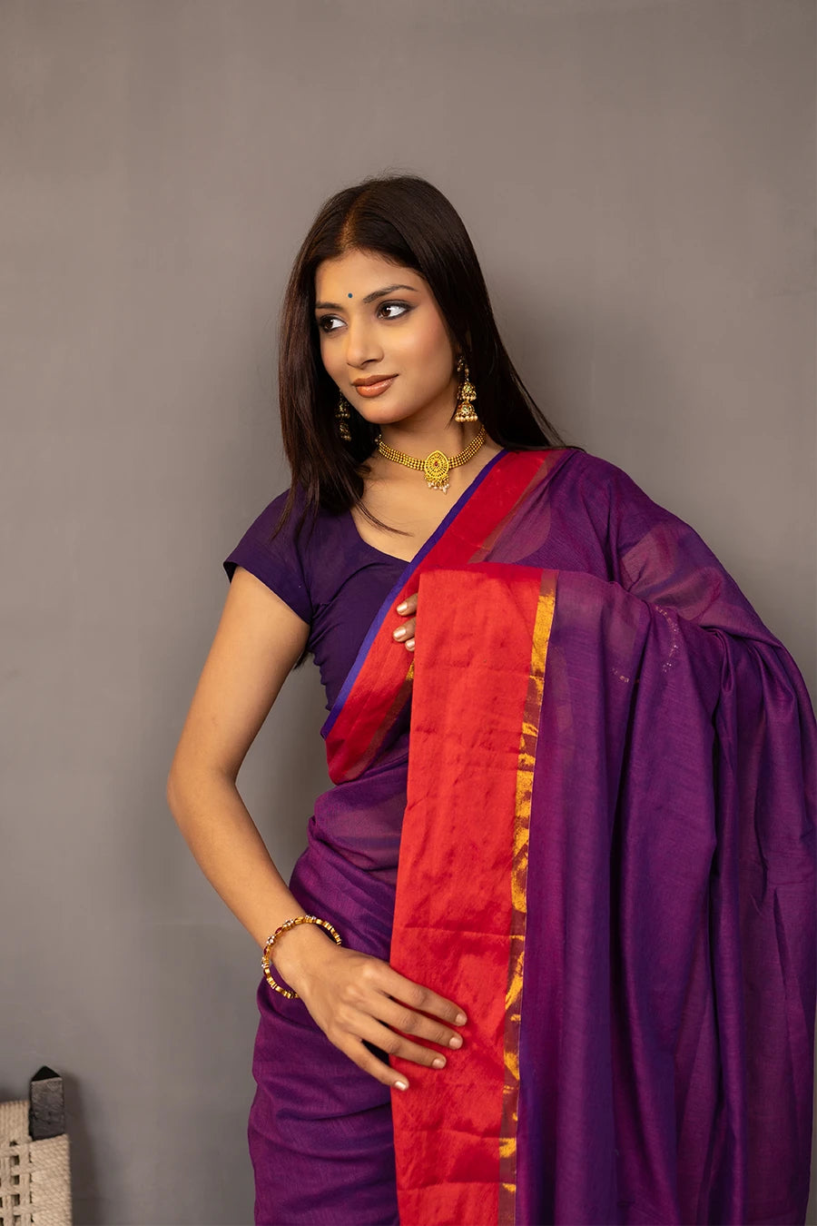 Woman wearing a purple saree with red border against a grey background