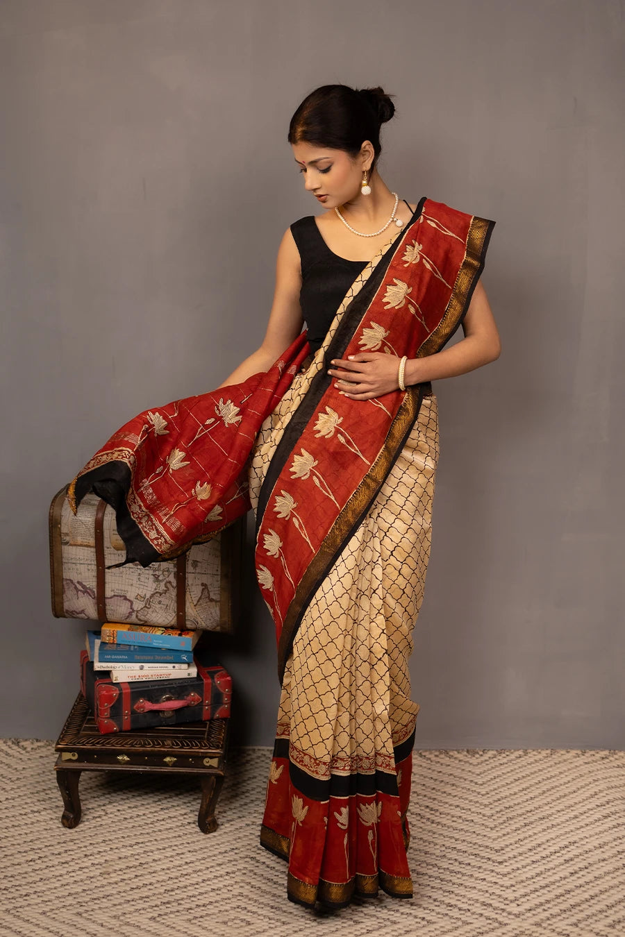 Woman wearing a traditional saree with a red border, standing next to a small table with books and a map.
