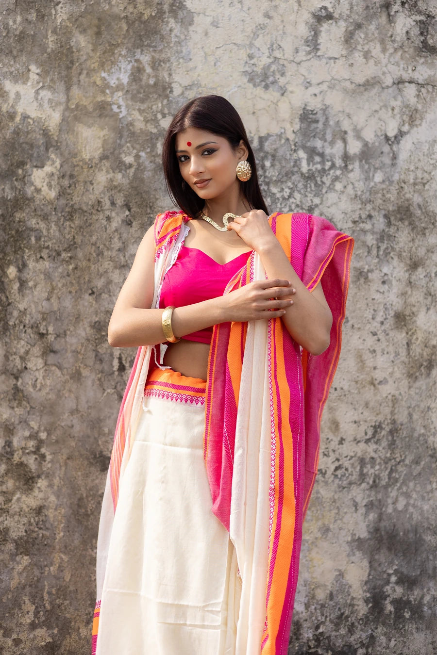 Woman wearing a traditional saree with pink and orange border against a textured wall.