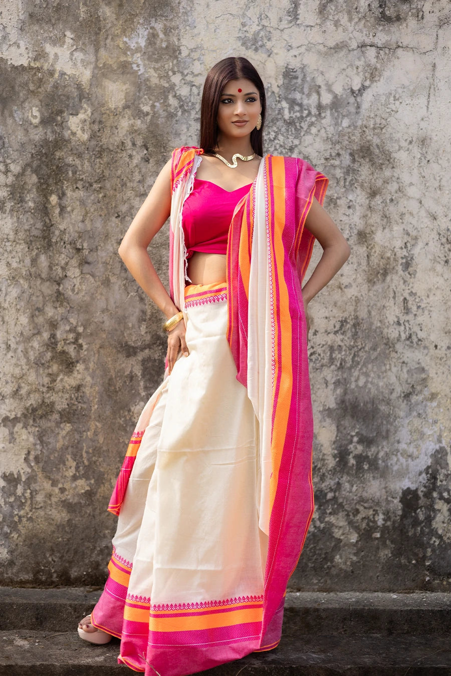 Woman wearing a traditional saree with pink and orange border against a textured wall.