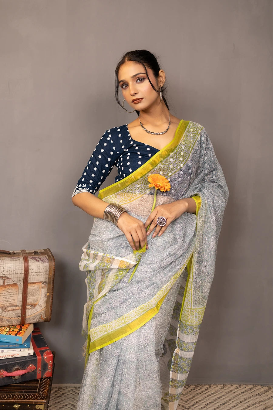 Woman in a saree standing next to a small table with books and boxes against a grey background