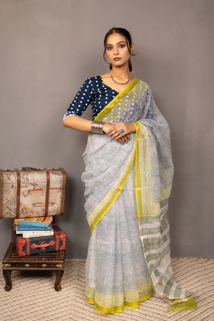 Woman in a saree standing next to a small table with books and boxes against a grey background