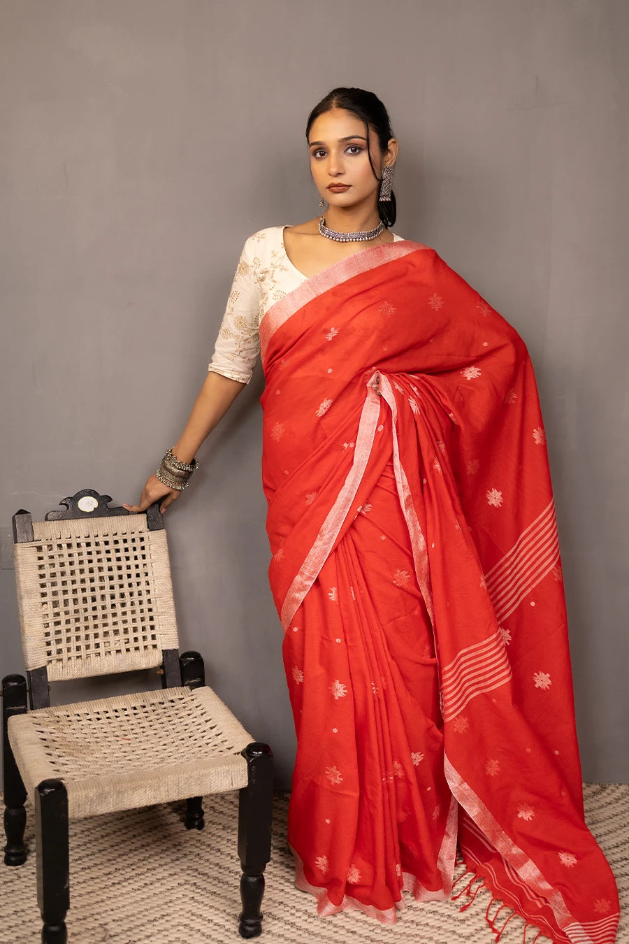 Woman in a red saree standing next to a chair against a grey wall