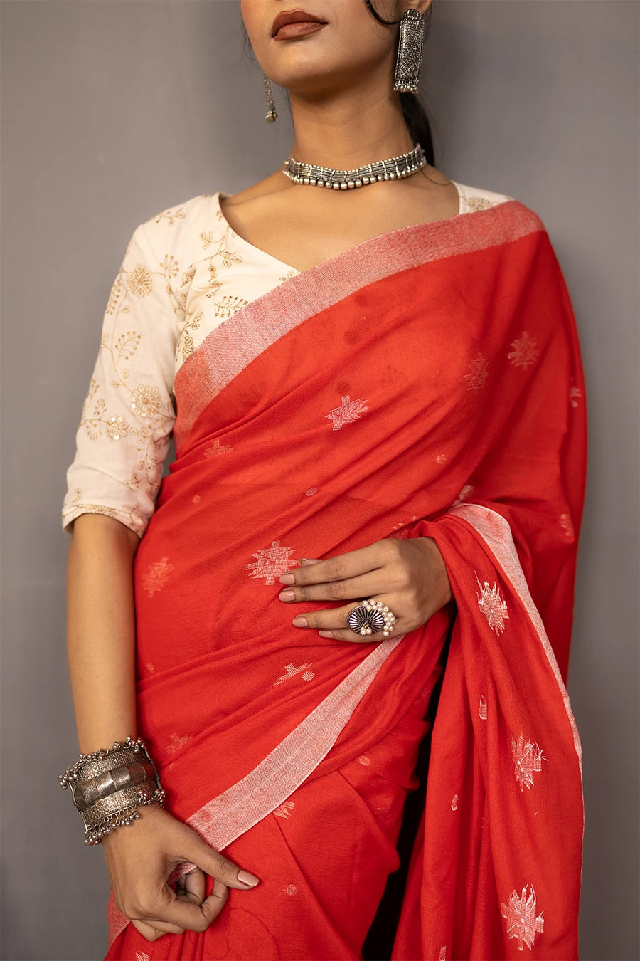 Woman in a red saree standing next to a chair against a grey wall