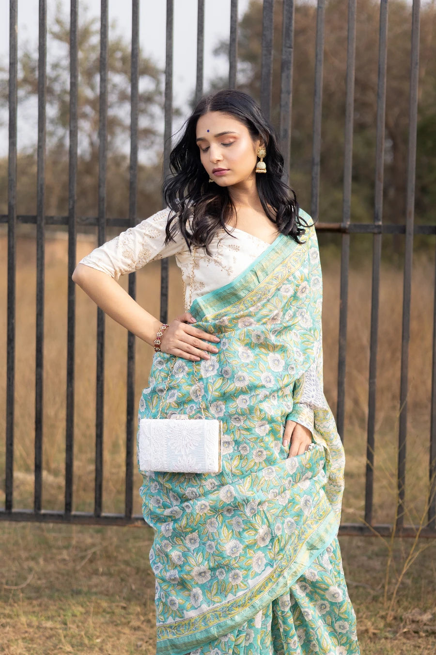 Woman wearing a green floral saree standing outdoors with a metal fence and grass in the background.