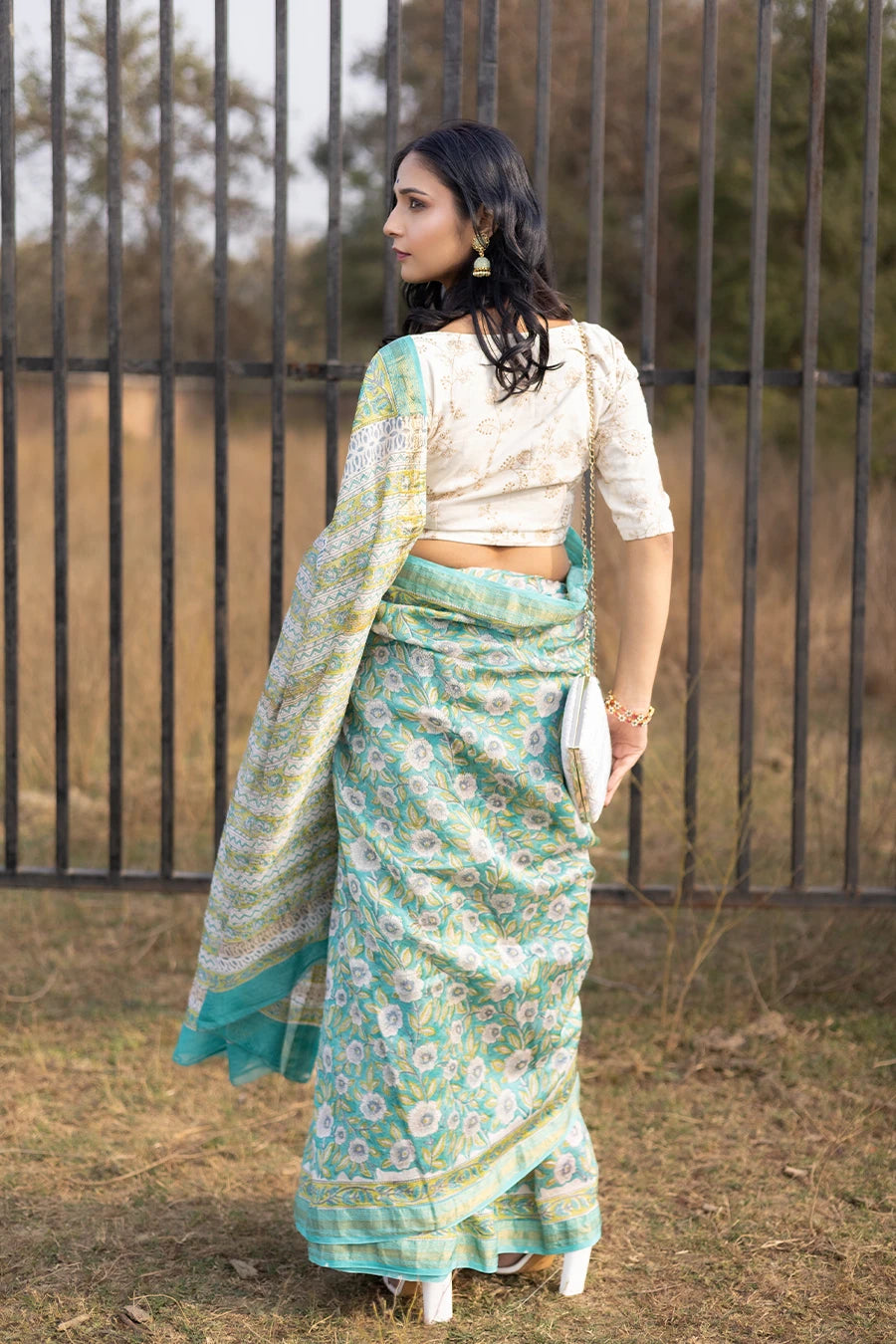 Woman wearing a green floral saree standing outdoors with a metal fence and grass in the background.