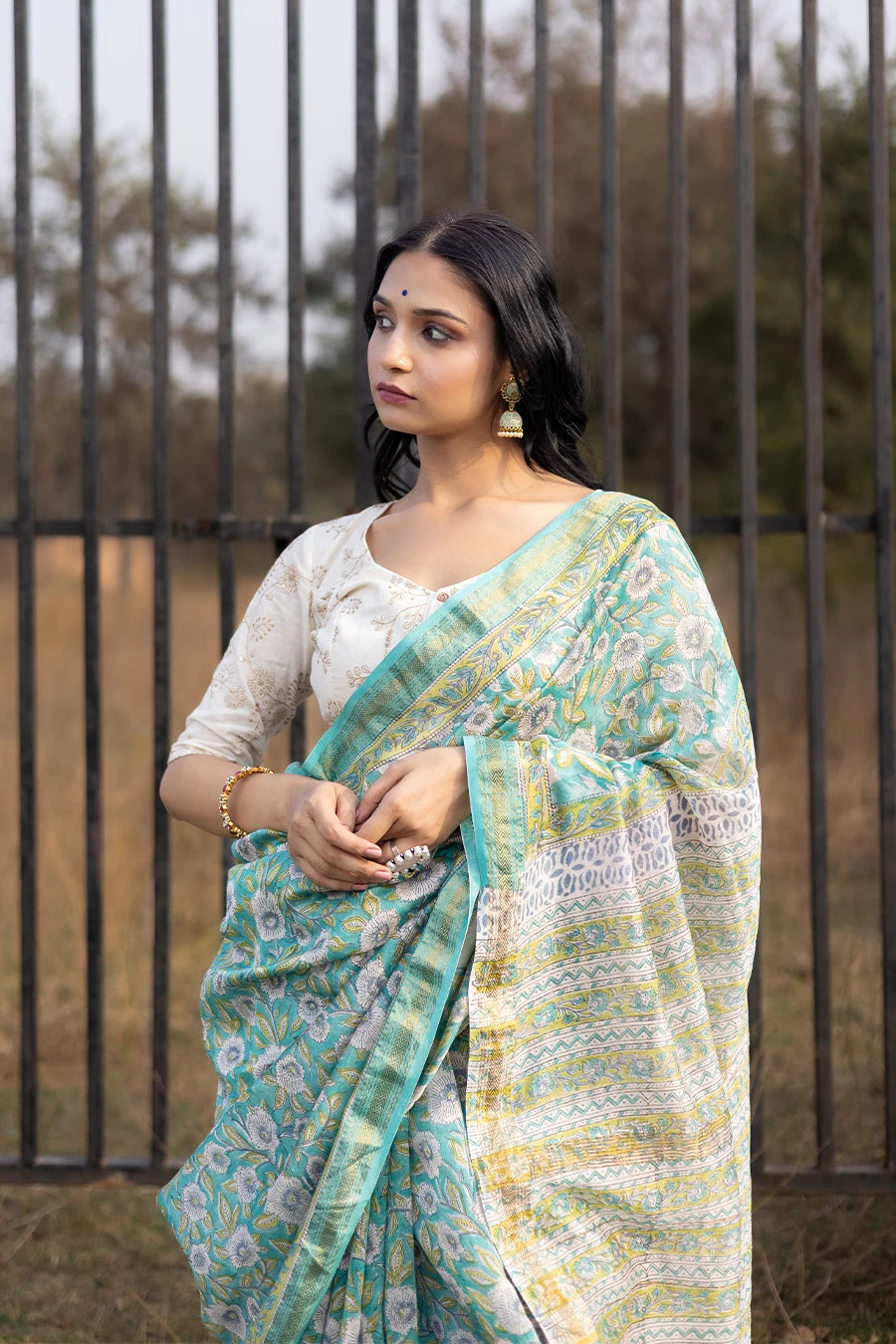 Woman wearing a green floral saree standing outdoors with a metal fence and grass in the background.
