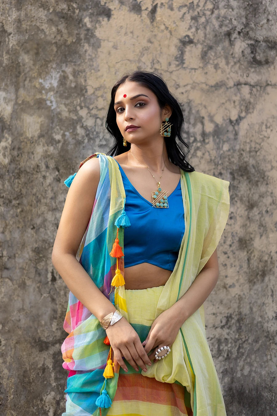 Woman wearing a colorful saree with a blue blouse against a textured wall.