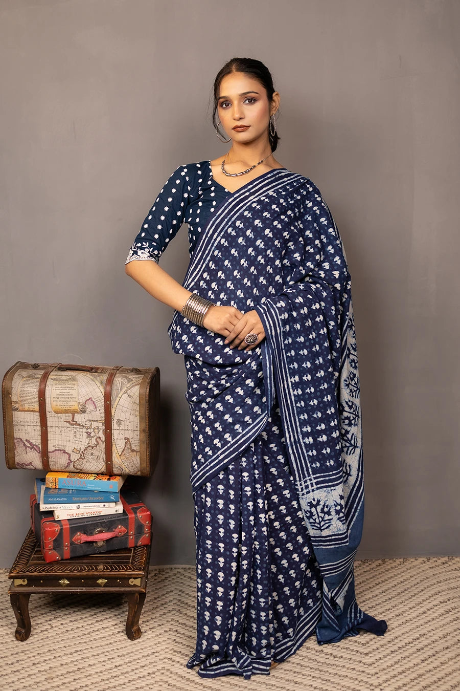 Woman wearing a blue saree with a pattern, standing next to a small table with books and a map.