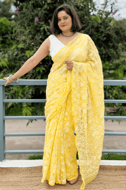 A woman wearing a bright yellow floral print saree with a white sleeveless blouse, standing outdoors in front of a green garden and a gray metal railing.