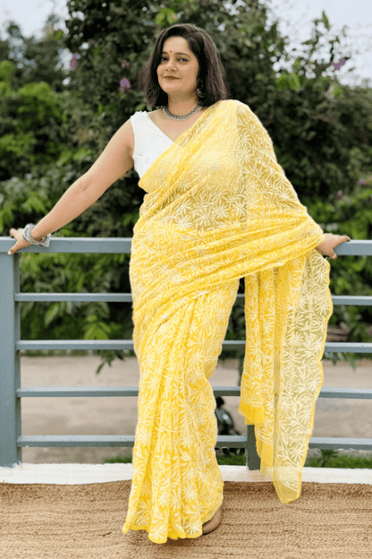 A woman wearing a bright yellow embroidered saree with a white sleeveless blouse, standing outdoors in front of a green garden and metal railing.