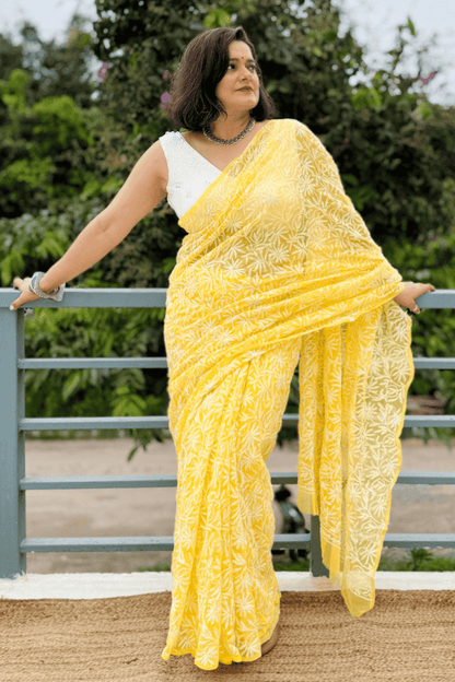 A woman wearing a bright yellow embroidered saree with a white sleeveless blouse, posing outdoors against a green garden background and a gray metal railing.
